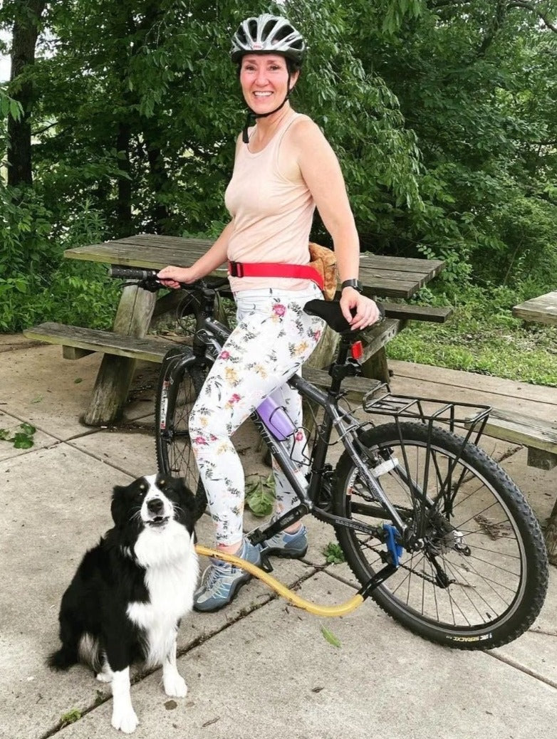 a rider in floral print leggings and a black and white Australian Shepherd, clipped to the BTL, turn backwards to look at the camera. This yellow Bike Tow Leash® uses the Chain Stay to attach to this bike
