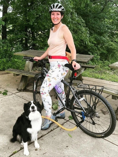 a rider standing on a bike with a black and white Border Collie, clipped to the BTL, turn backwards to look at the camera. This yellow Bike Tow Leash® uses the Chain Stay to attach to this bike.